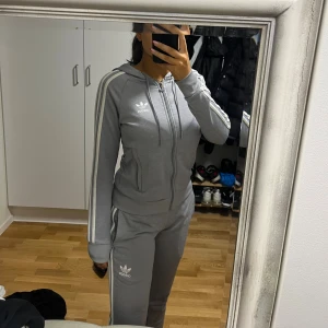 Grå Adidas hoodie med dragkedja - Säljer en grå hoodie från Adidas med klassiska vita ränder längs ärmarna och logga på bröstet. Tröjan har huva med snörning, dragkedja framtill och ribbade muddar. Perfekt för chill eller träning, snygg och sportig stil.