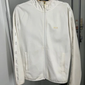 Vit zip-hoodie från BOSS - Vit hoodie från BOSS med dragkedja och huva. Tröjan har logotape längs ärmarna och diskret BOSS-tryck på bröstet i guld. Mjuk bomullskänsla och två fickor framtill. Perfekt för en clean och sportig look.