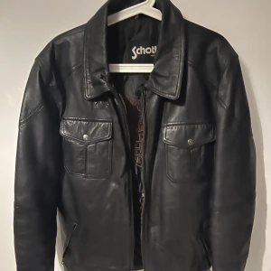 Svart skinnjacka från Schott NYC - Riktigt snygg svart skinnjacka från Schott NYC med klassisk krage, två stora bröstfickor med lock och knappar samt dragkedja framtill. Insidan har quiltat foder och broderad örnlogga. Perfekt för dig som vill ha en tidlös och cool look. 100% lammskinn