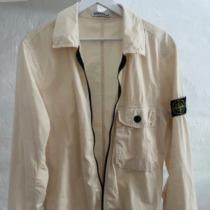 Beige overshirt från Stone Island - Snygg beige overshirt från Stone Island med klassisk logga på ärmen. Jackan har en svart dragkedja framtill, bröstficka med knapp och krage. Perfekt för lager-på-lager och stilren look.