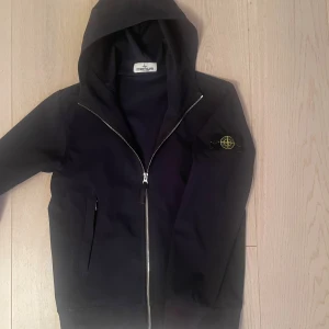 Svart vindjacka från Stone Island - Svart vindjacka från Stone Island med ikonisk patch på vänster ärm. Jackan har huva, hel dragkedja framtill och två sidofickor. Materialet är syntet och polyuretan, vilket gör den perfekt för blåsiga dagar. Snygg och stilren design som passar streetwear.