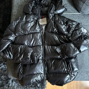 Svart Moncler pufferjacka med huva - Säljer en svart äkta  pufferjacka från Moncler med glansig finish och huva. Jackan har Moncler-logga på ärmen, dragkedja framtill och praktiska fickor. Fylld med gåsdun för extra värme och har coola detaljer som ribbade muddar och innerfoder med serietidningsmotiv.