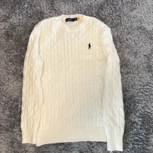 Vit stickad tröja Polo Ralph Lauren - Säljer en vit stickad tröja från Polo Ralph Lauren i storlek M. Tröjan har klassiskt kabelstickat mönster, rund hals och långärmade ribbade muddar. På bröstet finns den ikoniska svarta Polo-loggan broderad. Perfekt för dig som gillar stilren och tidlös design.
