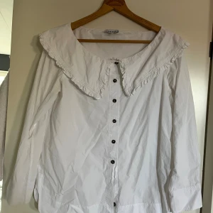 Vit skjorta med volangkrage från Ganni - Supersnygg vit skjorta från Ganni med stor volangkrage och bruna knappar framtill. Skjortan har långa ärmar och är gjord i mjuk bomull. Perfekt för dig som gillar statementkragar och vill ha en trendig look.