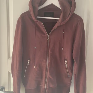 Vinröd hoodie från limitato - Säljer en vinröd hoodie från limitato med dragkedja framtill och två fickor med dragkedja. Jackan har huva med snörning och ett stort tryck på ryggen med motiv av bil och flygplan. Perfekt för dig som gillar streetwear.