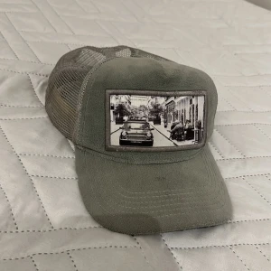Grå meshkeps med bilmotiv - Säljer en grå keps med mesh på sidorna och baksidan för extra ventilation. Framtill finns en patch med ett svartvitt motiv av en bil på en stadsgata. Kepsen har böjd skärm och justerbar passform bak.