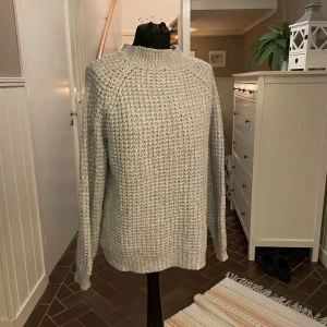 Grå stickad tröja - Säljer en grå stickad tröja med ribbad hals, ärmslut och nederkant. Tröjan har en klassisk rund hals och långa ärmar, perfekt för att hålla värmen under kyliga dagar. Enkel och stilren design som funkar till många olika outfits.