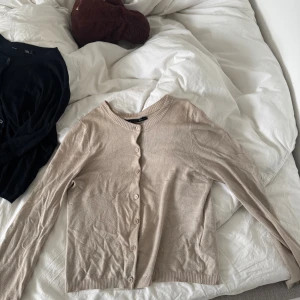 Beige och svart kofta från 157 - Två klassiska koftor från 157 i storlek S, en i beige och en i svart. Båda har rund halsringning, långa ärmar och knäppning framtill med matchande knappar. Perfekta att slänga över en topp eller klänning. Materialet känns mjukt och lätt.