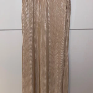 Champagnefärgad glittrig långklänning - Säljer en långklänning i champagnefärg med glittrigt, plisserat tyg och smala axelband. Klänningen har en elegant v-ringning och skimrande finish som fångar ljuset. 