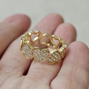 Guldring med glittriga hjärtan - Superfin guldfärgad ring med flera hjärtan runt hela bandet, där varje hjärta är täckt av små glittrande stenar. Perfekt för dig som älskar detaljer och vill ha något som sticker ut. Hjärtformen ger en söt och trendig vibe, och den glittriga ytan gör ringen extra iögonfallande.