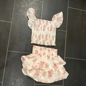 Vit blommig volangkjol från Kappahl och ett vit blommigt linne med ros blommor  - Supersöt kort volangkjol från Kappahl i vitt med rosa blommigt mönster. Både kjolen och linnet är väldigt stretchiga och sköna att ha på. Har andvände några gånger men fortfarande väldigt fin och somrig.😊