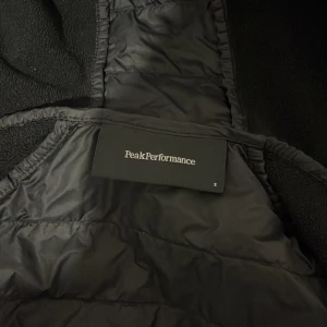 Peak Performance jacka  - Svart vindjacka från Peak Performance med huva och dragkedja framtill. Jackan har quiltad front och släta ärmar, samt logotyp på bröstet och ärmen. Materialet är syntet och polyester, perfekt för blåsiga dagar. Snygg och sportig look med normal passform.