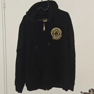 Svart hoodie med gul brodyr - Svart hoodie från med gul broderad Medusa-logga och texten på bröstet och ryggen. Tröjan har huva med snörning, dragkedja framtill med V-detalj och långa ärmar. Perfekt för dig som gillar exklusiv streetwear.