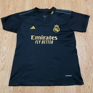 Real Madrid Fotbollströja barn - Svart Real Madrid fotbollströja från Adidas med guldiga detaljer, klubbmärke och sponsortryck på bröstet. Nummer 7 och VINI JR. på ryggen. Tillverkad i lätt och ventilerande Aeroready-material, perfekt för träning eller match.