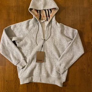 Grå hoodie från Burberry med dragkedja - Snygg grå hoodie från Burberry med klassiskt rutigt foder i huvan och broderad logga på bröstet. Tröjan har dragkedja framtill, snörning i huvan och två fickor. Perfekt för dig som vill ha en stilren och exklusiv look. Står XL men sitter mer som typ S