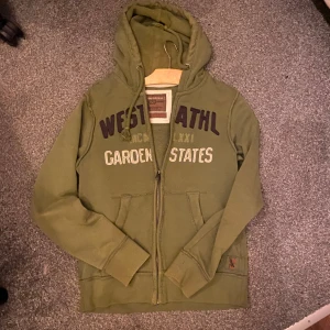 Olivgrön hoodie från Angelo Litrico - Olivgrön hoodie med dragkedja från Angelo Litrico. Tröjan har broderad text på bröstet, huva med snörning och två fickor framtill. Perfekt för en avslappnad stil och enkel att matcha med jeans eller joggers.