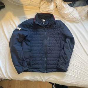 Mörkblå Helly Hansen Jacka - Mörkblå vinter/höst jagka från Helly Hansen med  dragkedja och ståkrage med röd detalj. Jackan har vita HH-loggor på ärmar och rygg, samt praktiska fickor med dragkedja. Perfekt för kyliga dagar och snygg till jeans. Väldigt enkel att göra en stilig outfit med. Funkar för alla tillfällen, om du har några frågor eller funderingar är det bara att skriva!