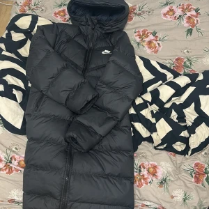 Svart lång pufferjacka från Nike - Säljer en svart lång pufferjacka från Nike med huva och dragkedja hela vägen. Köptes för 3500 ifrån Nike, har kvar taggarna nånstans och kvitto i mail, skicket är 10/10 Inga skador, eller fläck