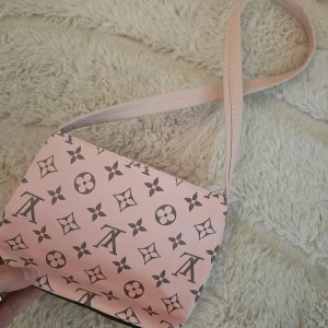 Rosa axelväska - Snygg rosa axelväska med klassiskt monogrammönster i svart. Väskan har en enkel, rektangulär form och ett brett axelband. Materialet är slätt och känns som syntet/skinn. Perfekt storlek för det viktigaste och ger en lyxig vibe till din outfit.