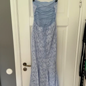Ljusblå långklänning med snörning - Superfin ljusblå långklänning med blommigt mönster och snygg snörning i ryggen. Klänningen har en elegant, figurnära passform och är ärmlös. Perfekt för sommarens speciella tillfällen. Materialet känns lätt och luftigt.