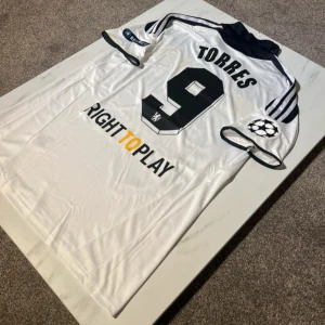Chelsea Torres #9 Adidas fotbollströja - Chelsea bortatröja med Torres #9 på ryggen, Adidas-märke och Champions League-patch på ärmen. Tröjan är vit med svart och gul panel över bröstet, klassisk krage och tre ränder på axlarna. Materialet är lätt och andas, perfekt för match eller samling.