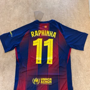 FC Barcelona Raphinha 11 matchtröja Nike - Säljer en officiell FC Barcelona matchtröja med Raphinha 11-tryck. Tröjan är kortärmad, tillverkad i lätt funktionsmaterial och har klassiska blå och röda ränder, klubbmärke, Nike-logga och sponsorer i gult. Perfekt för dig som älskar Barca och vill sticka ut på läktaren.