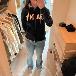 Mörkblå hoodie från GANT med dragkedja - Snygg mörkblå hoodie från GANT med vit logga framtill, huva med vita snören och praktisk dragkedja. Tröjan har två fickor fram och är tillverkad i mjuk bomullsmix. Perfekt för chill dagar och enkel att matcha med jeans eller joggers.
