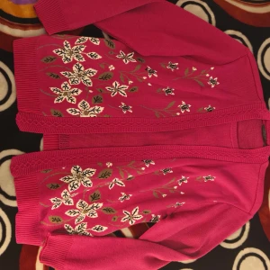 Rosa blommig kofta från Forever - Säljer en snygg rosa kofta från Forever by Michael Gold i storlek L/XL. Koftan har ett blommigt mönster i vitt, beige och grönt framtill, ribbade muddar och dekorativ kant. Materialet är stickat och känns mjukt mot huden. Perfekt för dig som gillar färg och detaljer!