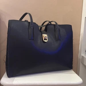 Svart väska, med guld detaljer - Snygg svart väska i skinn med stilrena raka linjer och rejäla handtag. Väskan har en guldfärgad låsdetalj framtill och ett rymligt fack med dragkedja på insidan. Perfekt för laptop och allt du behöver under dagen.