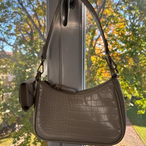 Beige axelväska från Ideal of Sweden - Snygg beige axelväska med krokodilpräglad yta från Ideal of Sweden. Väskan har guldfärgade detaljer och en matchande liten plånbok som hänger på sidan. Stängs med dragkedja och har stilren, modern form. 