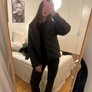 Svart quiltad jacka med knappar - Quiltad svart jacka med snyggt rutmönster och knappar framtill. Jackan har en loose passform och är långärmad, perfekt för dig som gillar en chill och stilren look. Materialet är syntet och jackan är lätt vadderad för extra komfort.