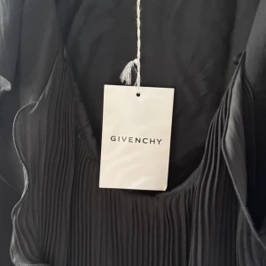 Svart plisserad klänning Givenchy - Supersnygg svart klänning från Givenchy med plisserade detaljer framtill och elegant volang vid urringningen. Klänningen har trekvartsärm och är tillverkad i 100% polyester. Perfekt för dig som vill ha en stilren och lyxig look.