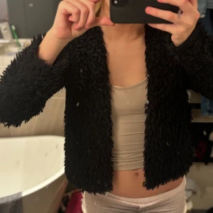 Svart pälsjacka med fluffig look - Säljer en svart pälsjacka med fluffig och mjuk känsla. Jackan har lång ärm och är i en kortare, normal passform. Perfekt för dig som vill ha en mysig och trendig jacka med cool textur. Enkel att matcha med olika outfits.