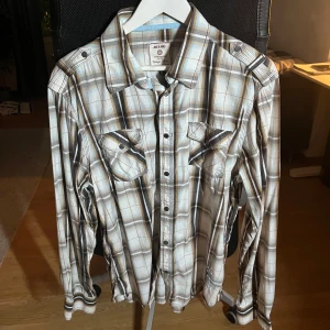 Rutig skjorta från Jack & Jones XL - Snygg rutig skjorta från Jack & Jones i beige, brunt och blått. Skjortan har klassisk krage, knappar framtill och två bröstfickor med lock. Långärmad modell i mjuk bomull, perfekt för en avslappnad stil.