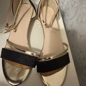 Svarta och guldiga sandaler med glitter storlek 38 - Snygga sandaler med svart glittrig ovandel och guldfärgade detaljer. Remmarna är i metallic guld och har spänne med liten dekoration. Klacken är låg och har en cool geometrisk guldlook. Perfekta för dig som vill sticka ut med stilrena detaljer.