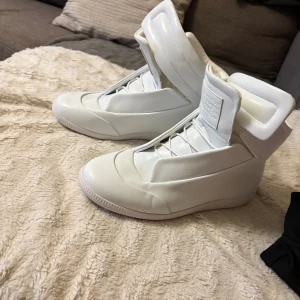 Vita futuristiska sneakers med hög plös - Säljer ett par vita sneakers från Maison Margiela med unik, futuristisk design och hög vadderad plös. Skorna har slät ovandel, diskret snörning och är tillverkade i skinn för en clean och modern look. Perfekta för dig som vill sticka ut med din stil.