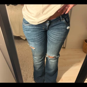 Blå bootcut jeans med slitningar - Snygga blå jeans med bootcut-modell och slitna detaljer vid knäna. Låg midja och klassisk femficksdesign. Jeansen har en cool tvätt och är perfekta för dig som gillar en avslappnad och trendig stil.