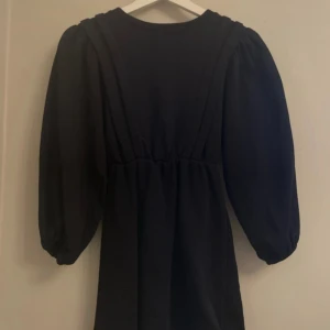 Svart klänning med puffärmar från LMTD - Svart klänning från LMTD med markerad midja och snygga puffärmar. Modellen är enkel men trendig, med rund halsringning och trekvartsärmar som ger en cool siluett. Perfekt för dig som gillar stilrena plagg med en twist.