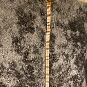 Burberry rutig bälte med guldspänne - Säljer ett smalt Burberry-bälte med klassiskt rutigt mönster i beige, svart och rött. Bältet har ett snyggt guldigt metallspänne med Burberrys ikoniska logga. Materialet är skinn och bältet har en stilren och modern look som lyfter din outfit. (110 cm) Man kan byta håll på den så den byter färg till svart