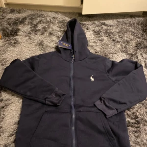 Marinblå hoodie från Polo Ralph Lauren - Snygg marinblå hoodie från Polo Ralph Lauren med dragkedja och huva. Tröjan har långa ärmar, två fickor framtill och den klassiska Polo-loggan broderad på bröstet. Tillverkad i mjuk bomull som är perfekt för chill dagar.