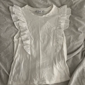 Vit volangtopp från MANGO - Supersöt vit topp från MNG med breda volanger i spets längs axlarna. Ärmlös modell i mjuk bomull, perfekt för en fräsch och stilren look. Volangdetaljerna ger toppen en romantisk touch och gör den extra speciell.