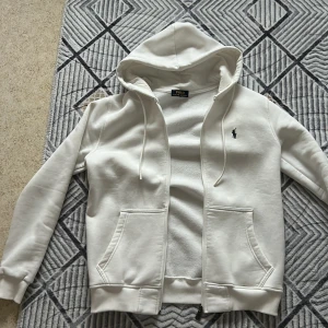 Vit hoodie från Polo Ralph Lauren - Vit hoodie från Polo Ralph Lauren i storlek S. Tröjan har huva med snörning, två fickor fram och en silverfärgad dragkedja med Polo-logga. Klassisk broderad logotyp på bröstet. Mjuk insida och ribbade muddar vid ärmslut och nederkant. Perfekt för chill dagar.