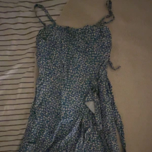 Blommig blå omlottklänning - Säljer en söt blå omlottklänning med vita blommor och smala axelband som knyts. Klänningen har en lätt och flowy känsla, perfekt för varma dagar. Den har en feminin passform och fina detaljer med knyt i midjan.