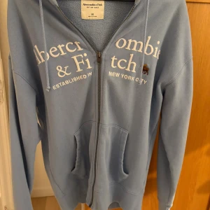 Blå hoodie från Abercrombie & Fitch XS - Säljer en chill blå hoodie från Abercrombie & Fitch i mjuk fleece. Tröjan har dragkedja, huva med snörning och stora fickor framtill. Vit broderad logga och moose-detalj på bröstet. Perfekt för dig som gillar relaxed fit och snygg streetstil. Har använt ca 3 gånger. Bra skick!!