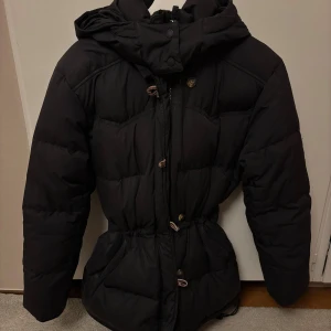 Svart dunjacka från Boomerang S - Svart dunjacka från Boomerang med huva. Jackan har både dragkedja och knappar framtill, samt detaljer med markerad snörning i midjan. Slitage och defekter på bilderna 4 & 5. Använt skick.