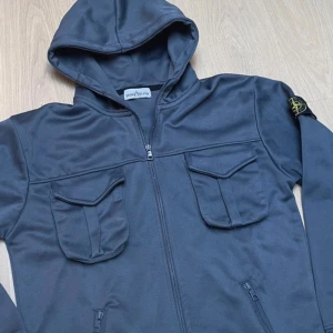 Mörkgrå hoodie från Stone Island - Snygg mörkgrå hoodie från Stone Island med huva, dragkedja och två stora fickor med lock framtill. Ikonisk patch på ärmen och detaljerade knappar med logga. Mjukt material och klassisk streetwear-vibe, perfekt för dig som gillar stilrena och funktionella plagg.