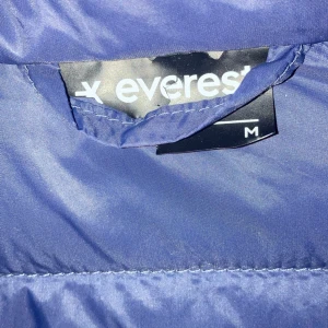 Mörkblå pufferjacka från Everest - Säljer en mörkblå pufferjacka från Everest i storlek M. Jackan har quiltad design, hög krage och dragkedja framtill. Perfekt för kalla dagar och har två sidofickor med dragkedja. Snygg och enkel att matcha med din stil.