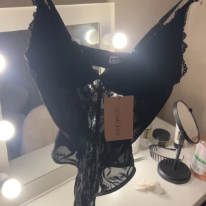 Svart spetsig body från SHEIN BAE - Säljer en svart body med transparent spets och fina detaljer från SHEIN BAE. Modellen har justerbara axelband, spetskant och en liten rosett framtill. Perfekt för dig som gillar edgy och snygga toppar med lite extra attityd.