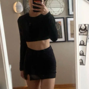 Svart croppad långärmad topp med öppen rygg och kjol med slits  - (Välj 3 plagg för 70kr - gäller alla plagg som kostar mellan 20-40kr) Säljer en svart croppad långärmad topp med öppen rygg och knytband och en kjol med slits och inbyggda shorts. Säljer som ett sett. 