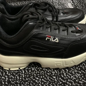 Svarta Fila sneakers med chunky sula - ’’INTE OG’’.                                                       Svarta Fila sneakers med vit chunky sula och broderad logga på sidan och hälen. Skorna har snörning och är tillverkade i syntetmaterial med vadderad insida för extra komfort. Perfekta för dig som gillar en sportig och trendig look.